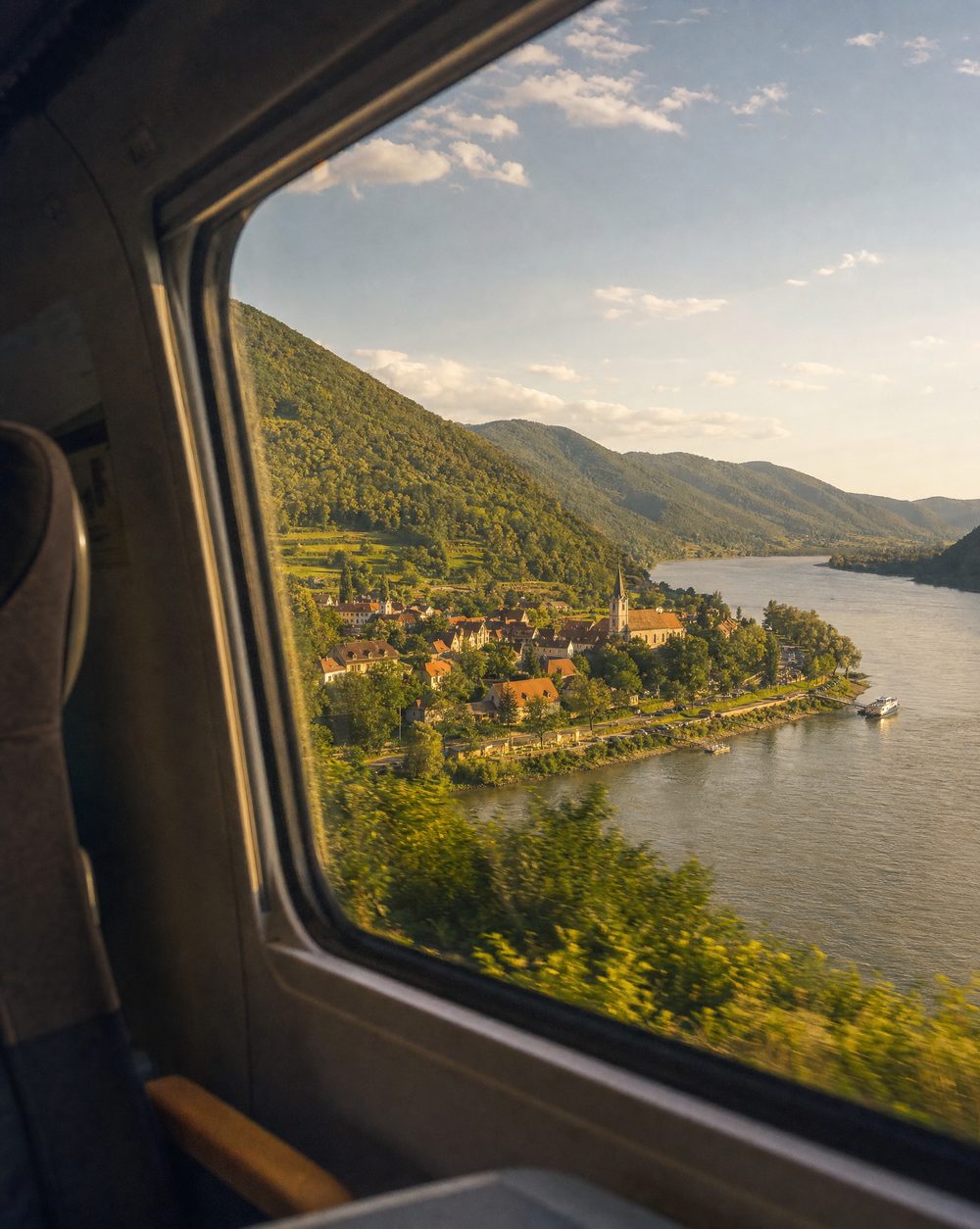 View from a train window of the Danube river bend, green hills, and a riverside village in golden afternoon light