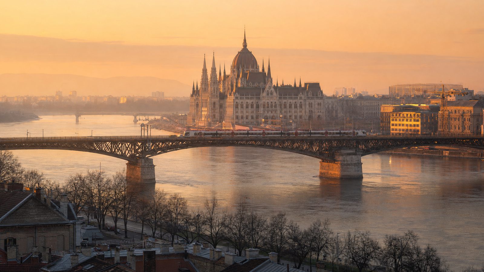 Golden-hour view across the Danube of the Hungarian Parliament Building in Budapest, with an intercity train crossing the bridge in the foreground